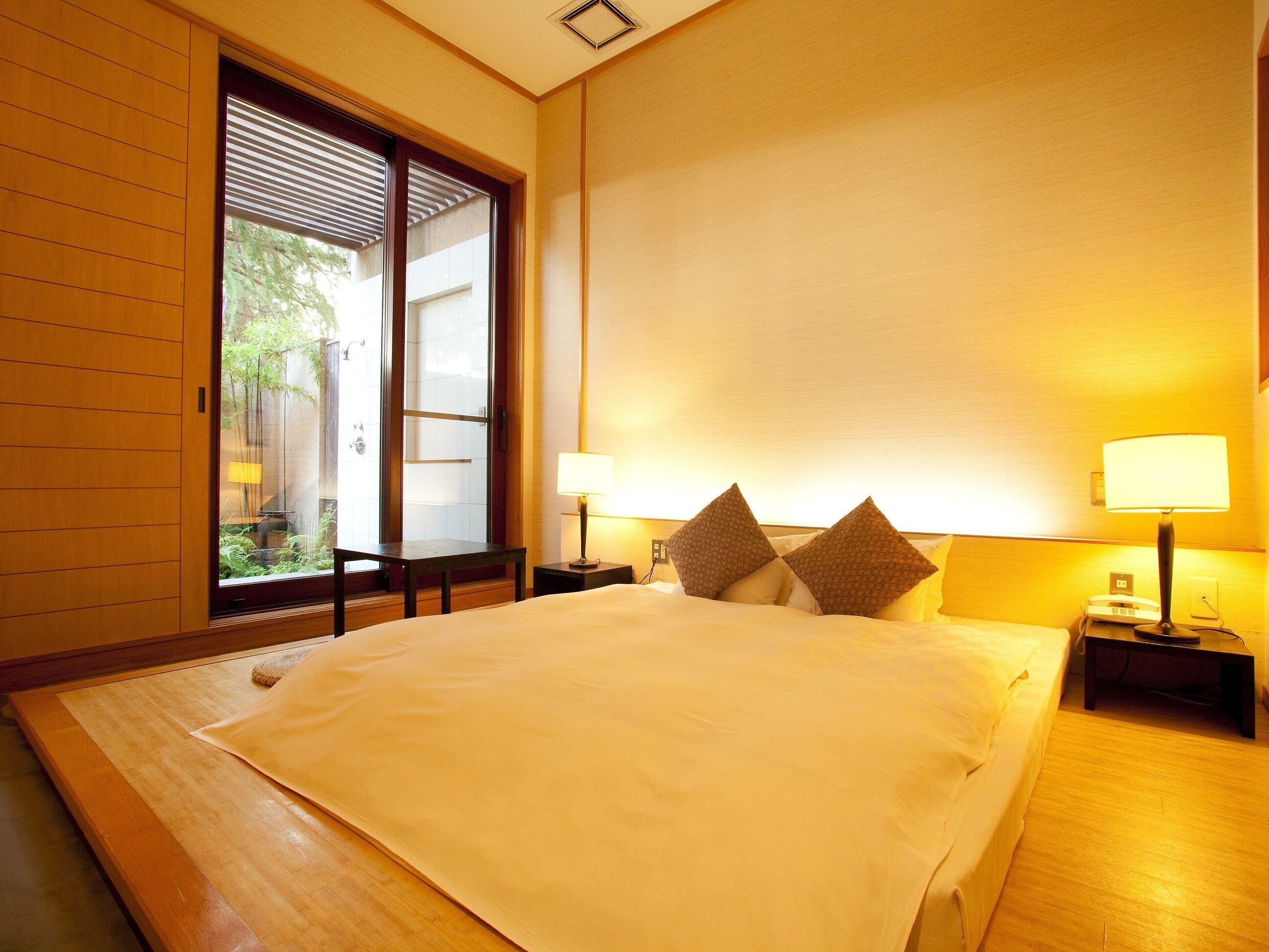 A hotel room with a large bed, two bedside lamps, and a view of greenery through a sliding door.