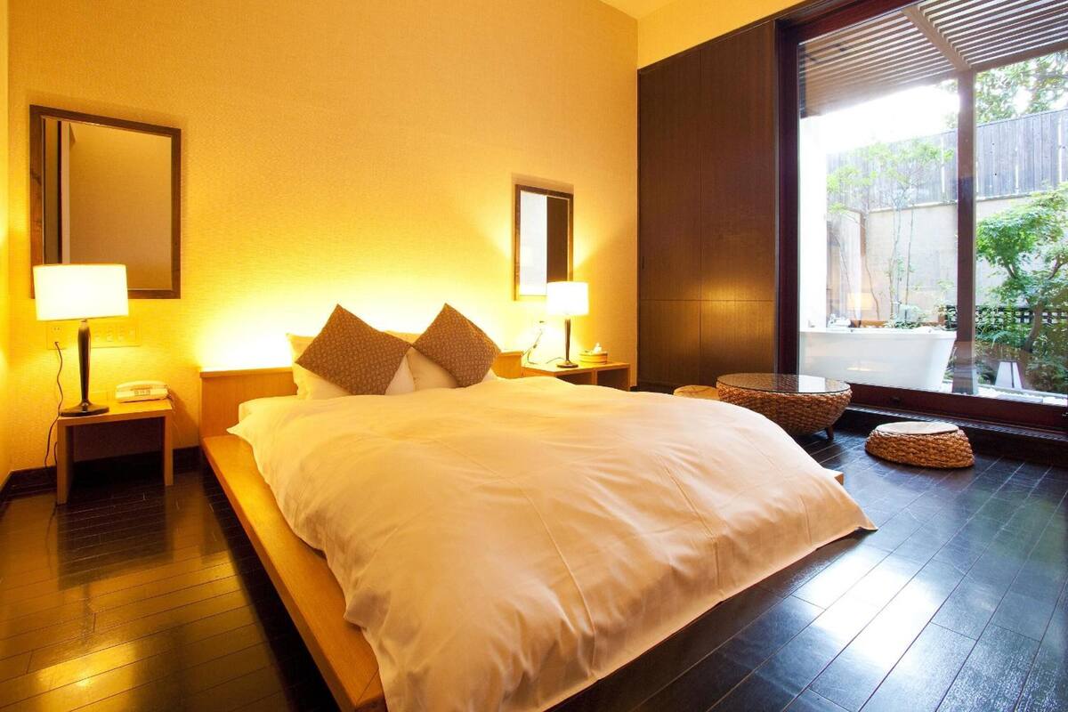 A hotel room with a large bed, two bedside tables with lamps, a large window with a view of greenery, and a dark wooden floor.