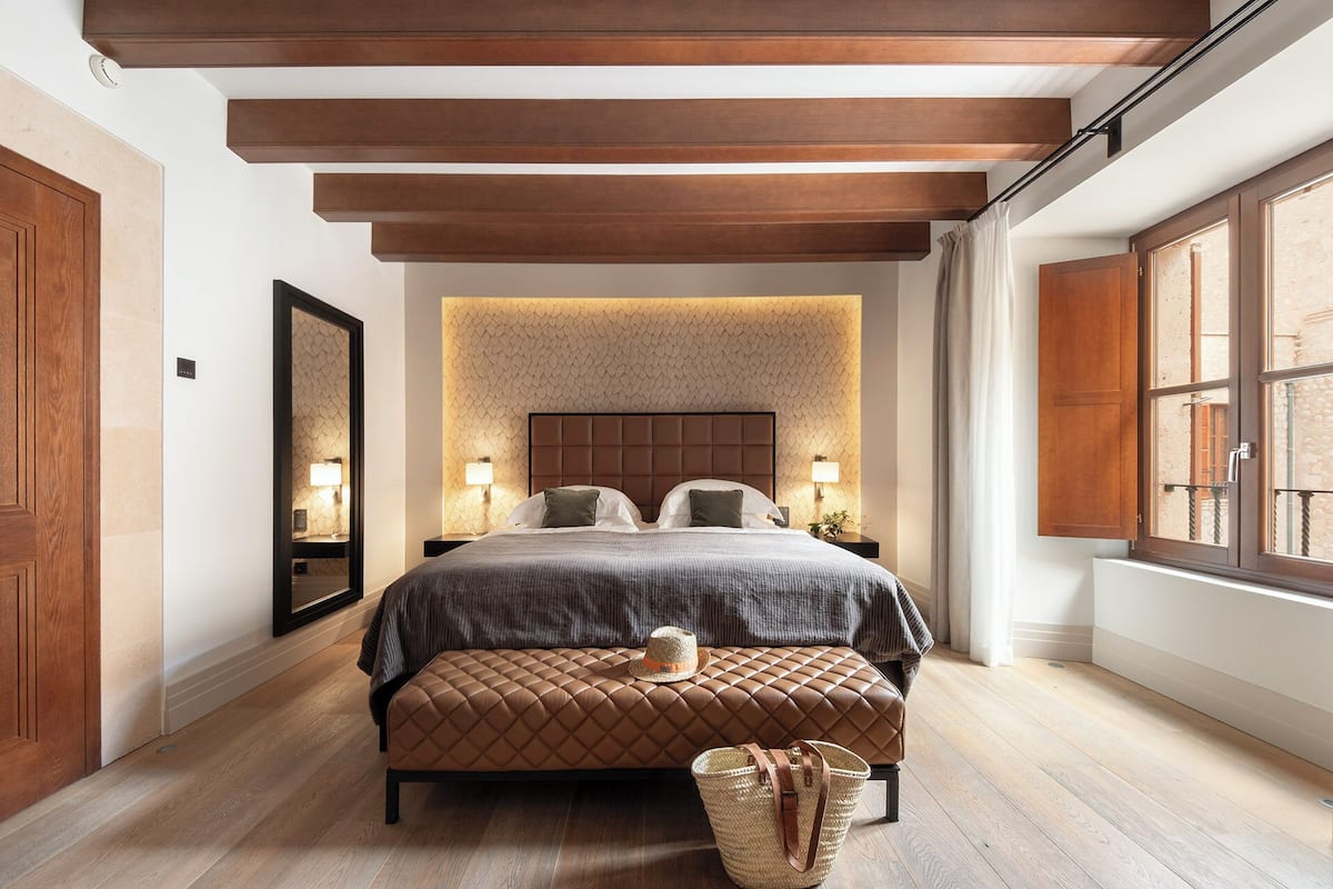 A bedroom with a large bed, a headboard with a textured wall behind it, a wooden bench, and a window with curtains.