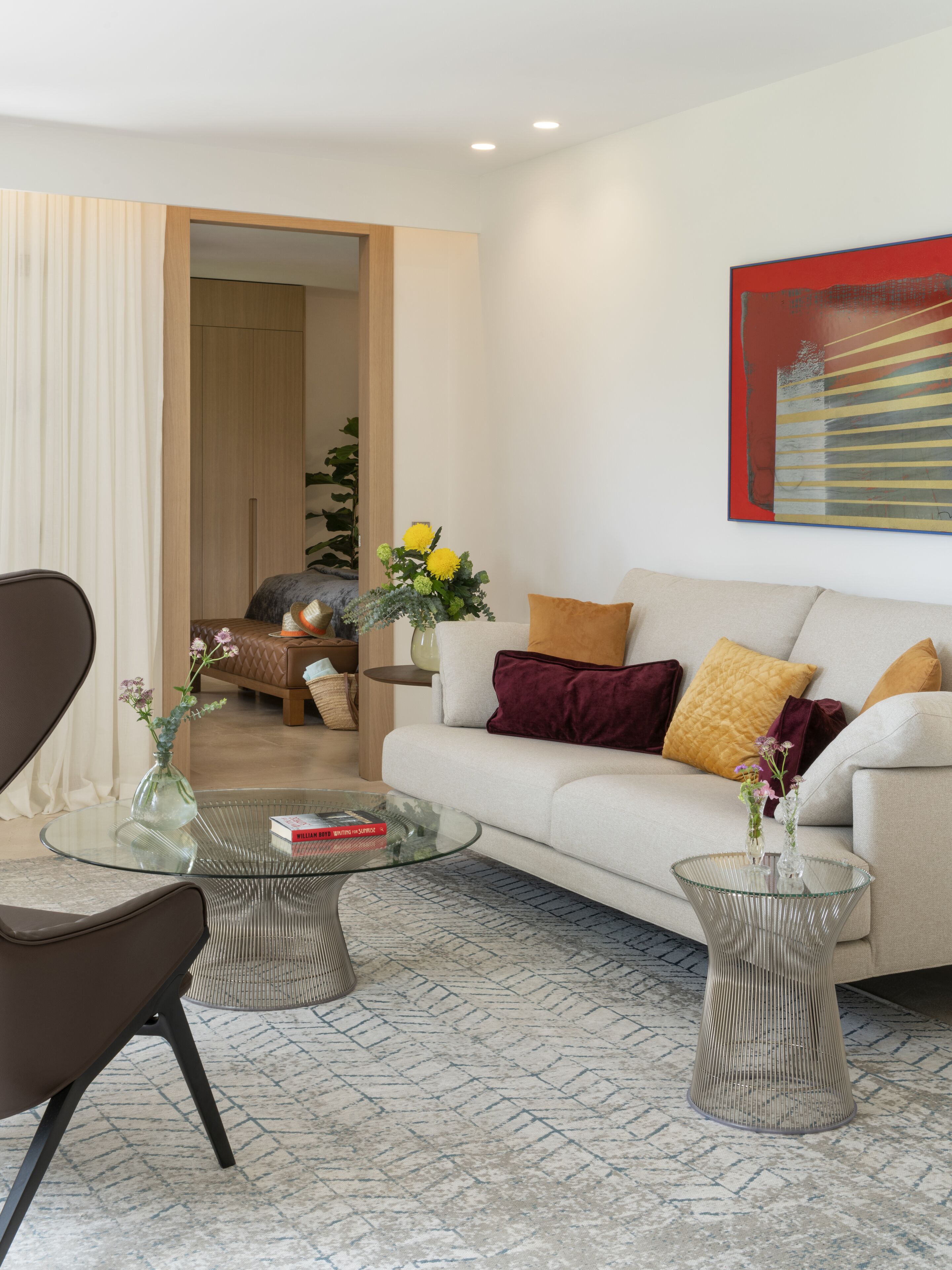 Una sala moderna con un sofá, dos mesas de centro de vidrio y una silla. Se ve una puerta que da a otra habitación y un cuadro abstracto de gran tamaño en la pared.