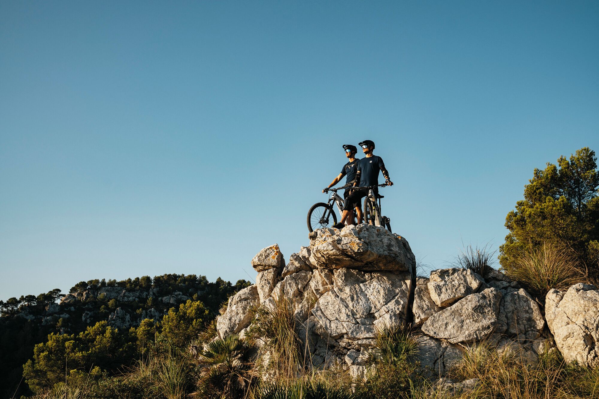 Two people standing on a rocky outcrop with bicycles, surrounded by trees and a clear sky.