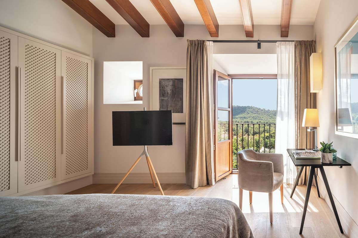 A hotel room with a bed, a desk with a lamp, a chair, a TV on a stand, and a balcony with a view of the hills.