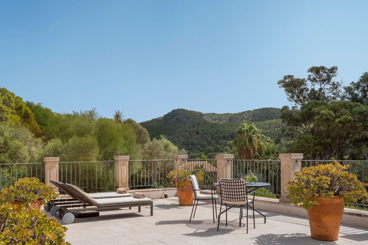 A terrace with a lounge chair, a table, and chairs, surrounded by potted plants and a view of a mountain.