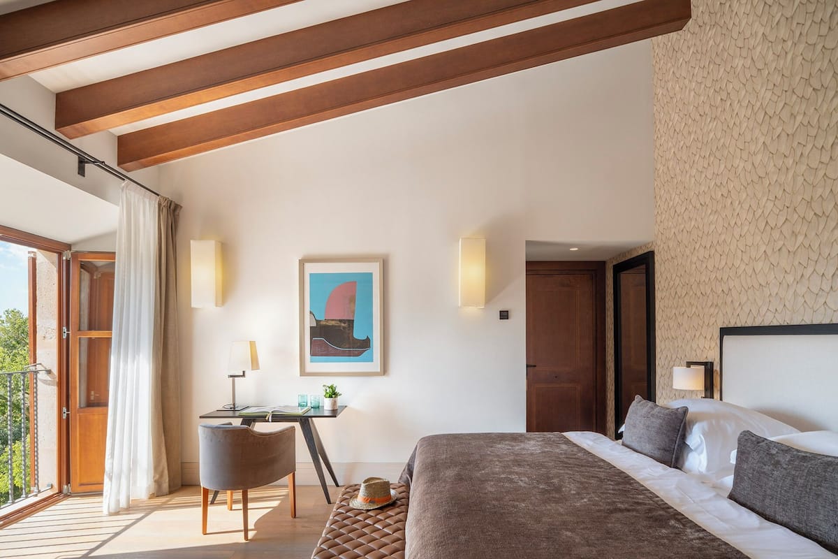 A modern bedroom with a large bed, a desk, and a chair. There is a balcony with a view of greenery.