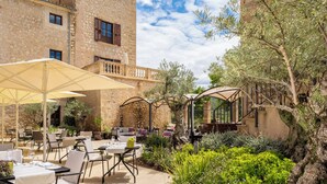 An outdoor patio with tables and chairs, surrounded by stone buildings and greenery.
