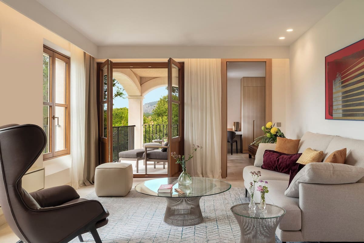 A modern living room with a glass coffee table, a sofa, and a chair. There is a balcony with a view of greenery.