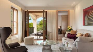 Un salon moderne avec un canapé, un fauteuil et une table basse en verre. Il y a un balcon avec du mobilier d’extérieur et une vue sur la verdure.