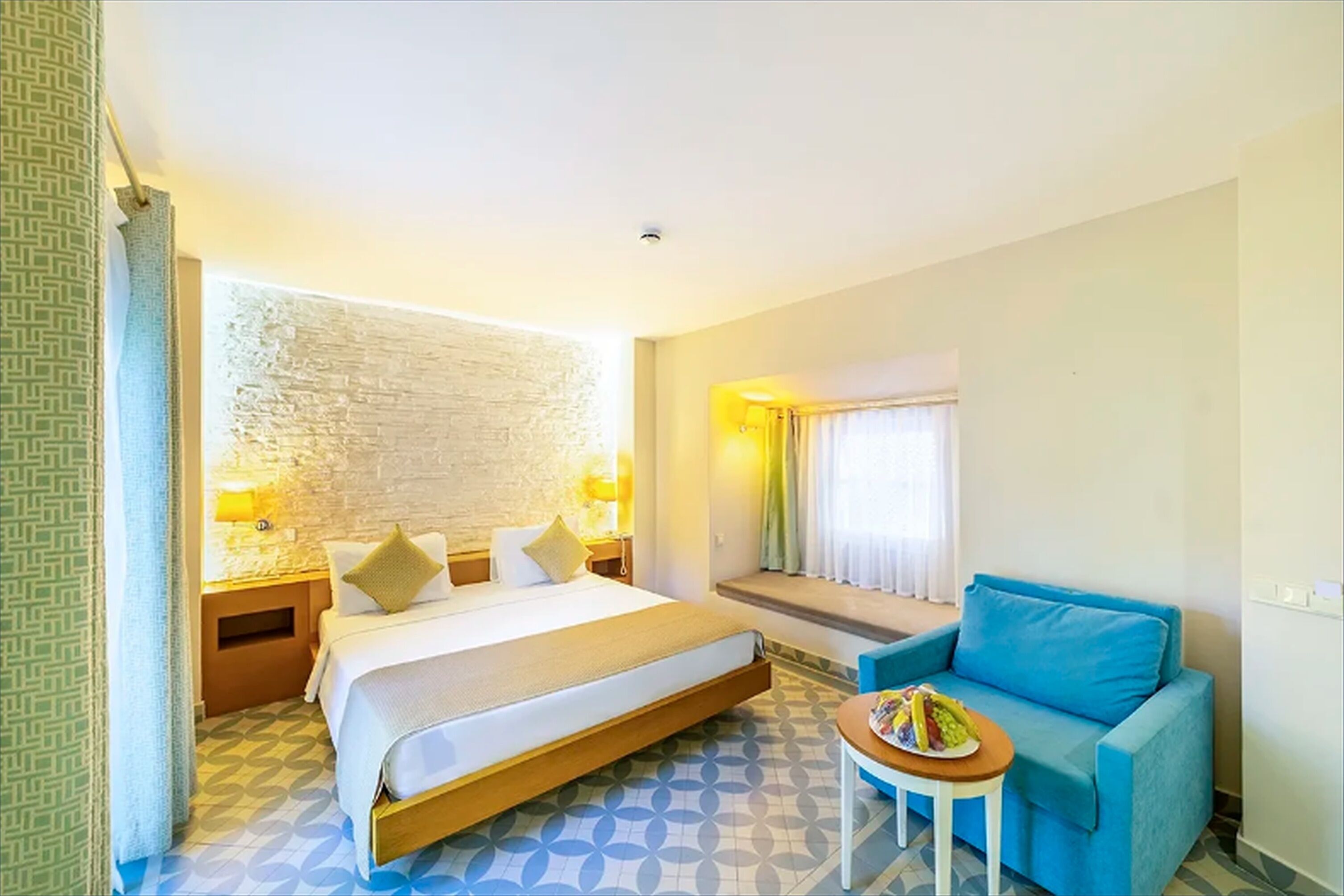 A hotel room with a bed, a blue sofa, a small table with fruit, a window with curtains, and a patterned floor.