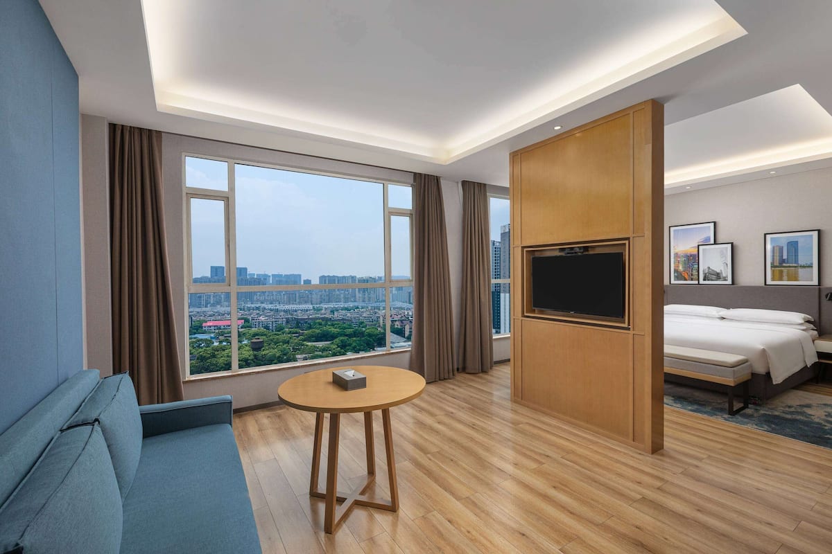 A hotel room with a large window, a sofa, a round table, a bed, a TV, and framed pictures on the wall.