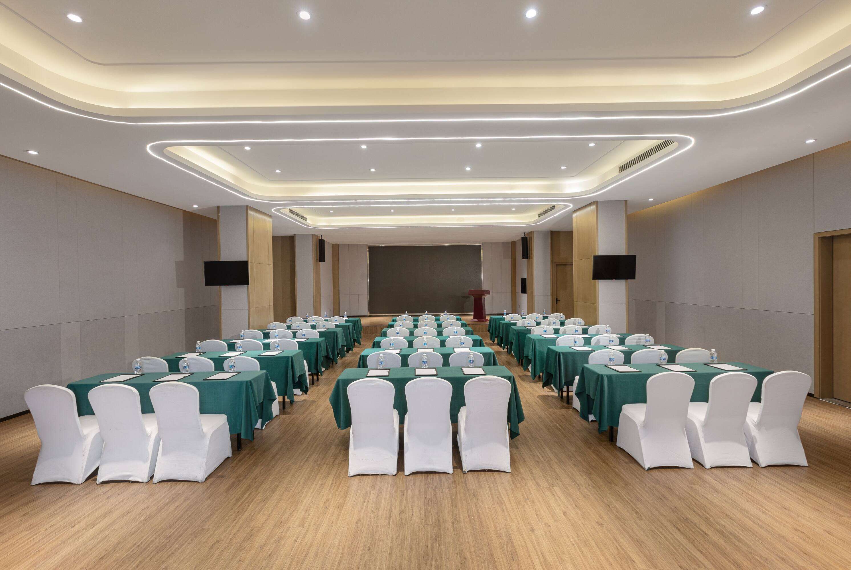Un salón de conferencias con filas de mesas y sillas, un atril y una pantalla grande al frente.