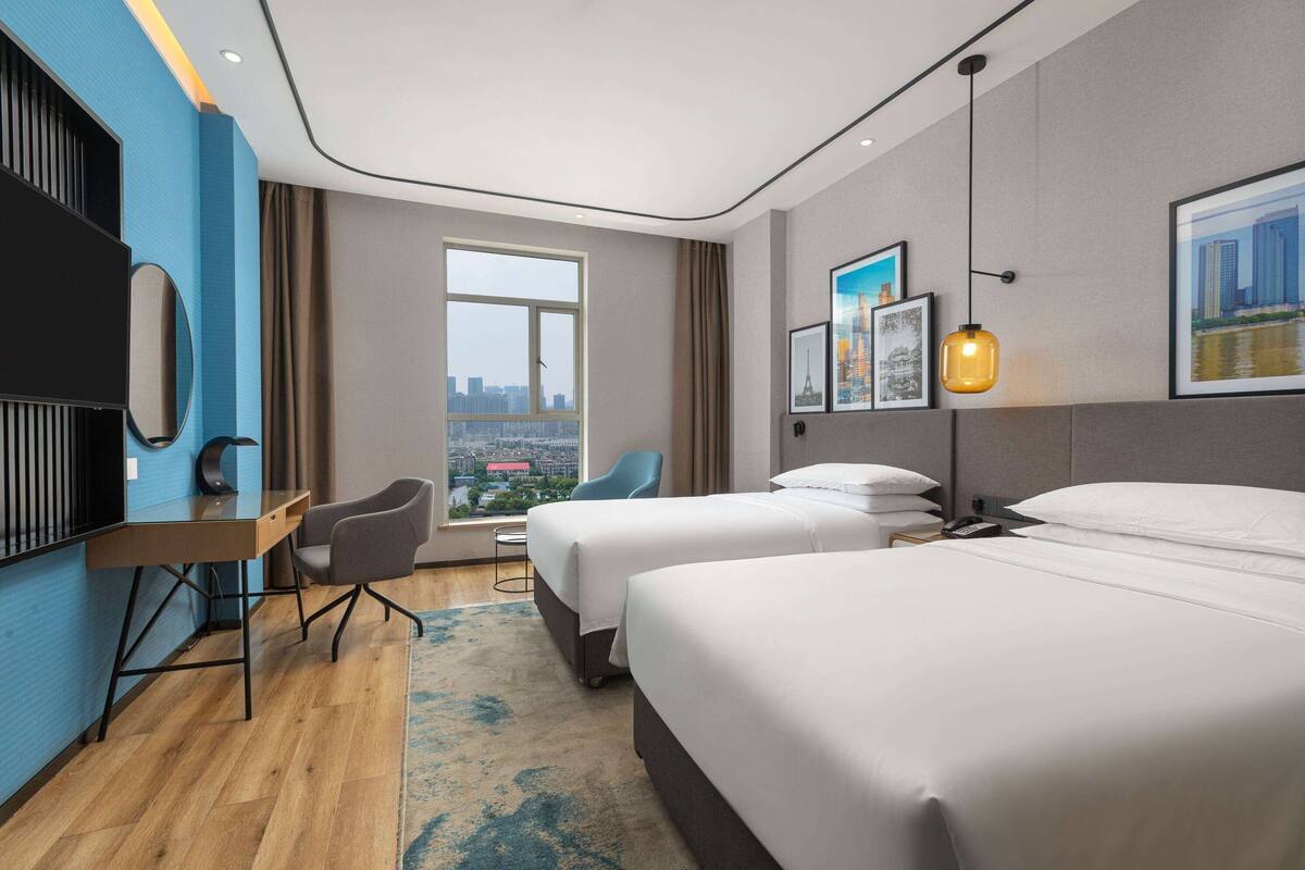 A hotel room with two beds, a desk, a chair, a large window with a city view, and artwork on the walls.