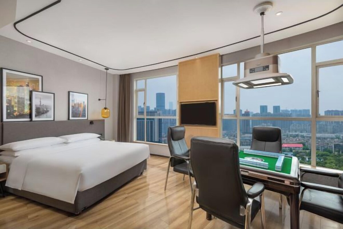 A modern hotel room with a large bed, a desk, and a chair, a city view through large windows, and a mounted TV.