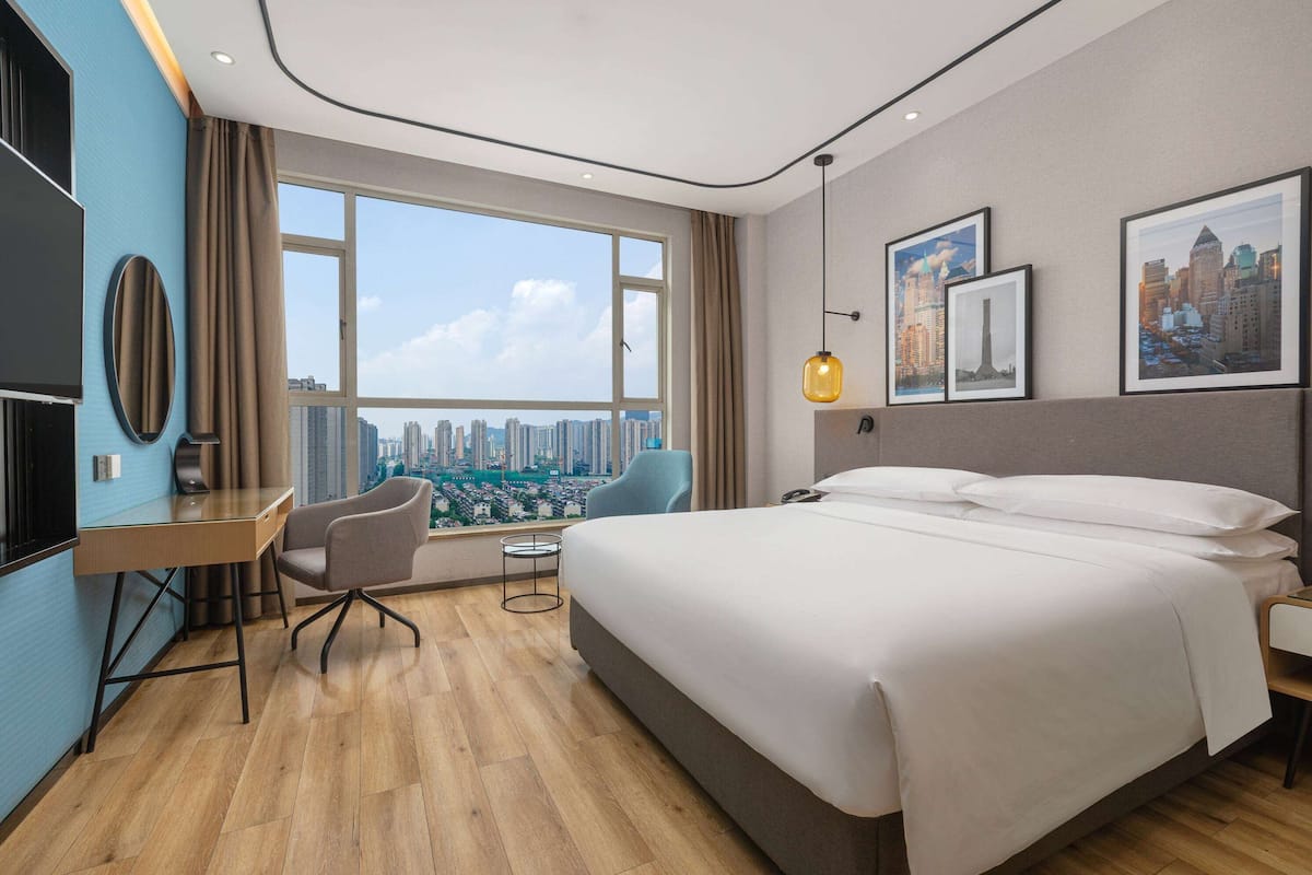A modern hotel room with a large bed, a desk, and a chair. There is a large window offering a city view.