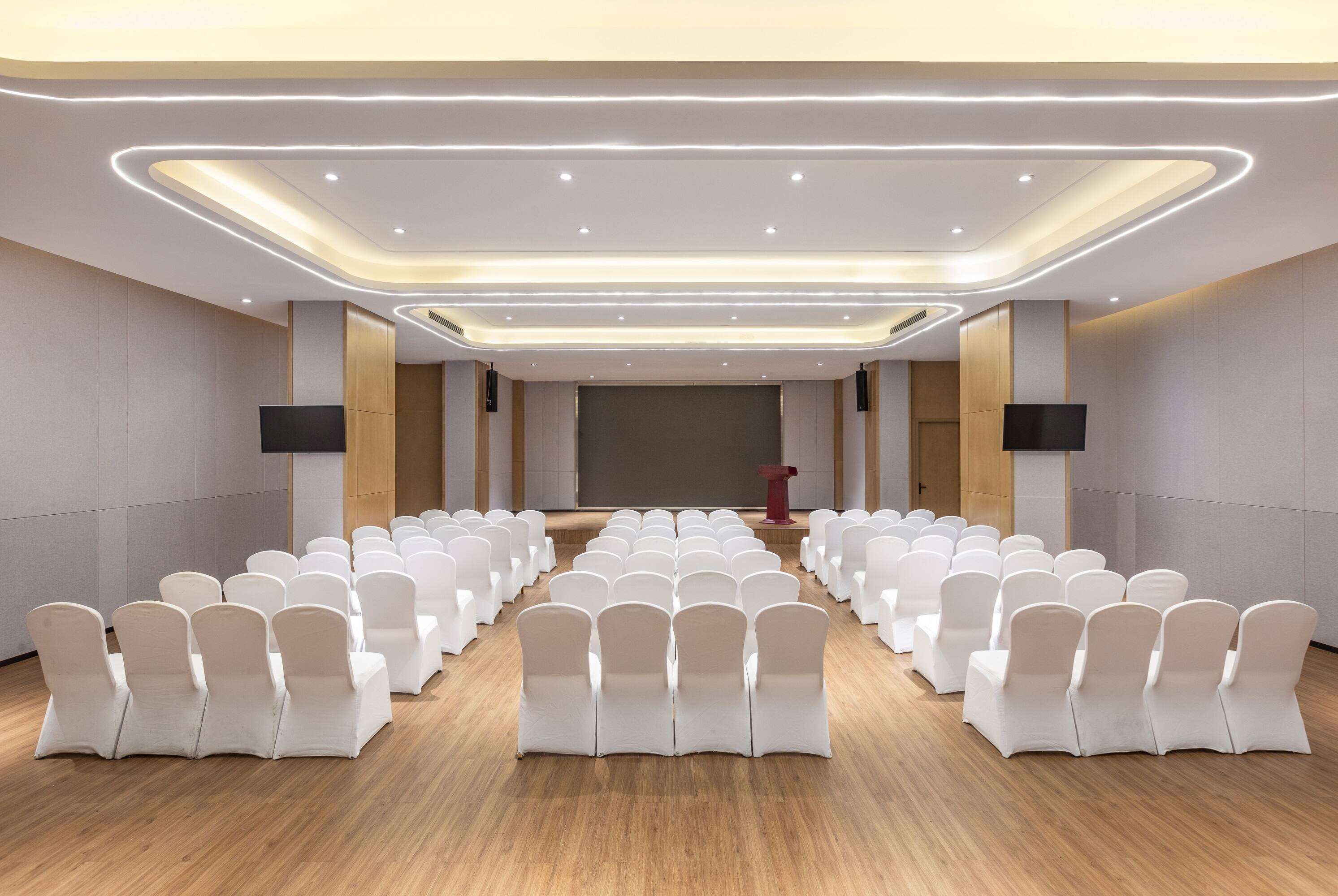 A conference room with rows of white chairs, a stage, and a modern ceiling design.
