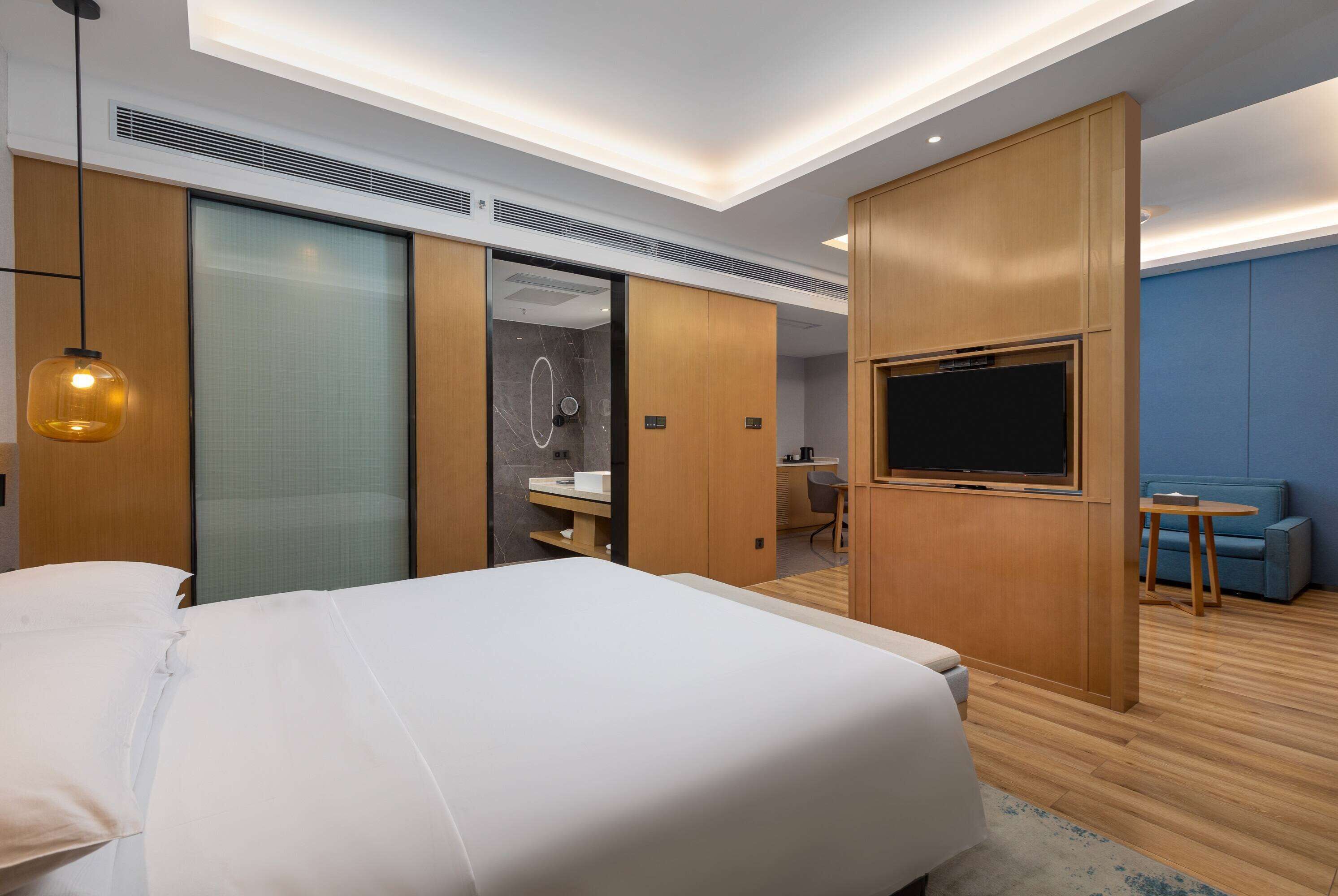 A modern hotel room with a large bed, a TV, a desk, and a seating area.
