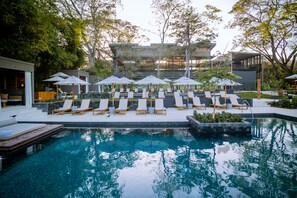 Zona de piscina con sillones reclinables y sombrillas, rodeada de vegetación y arquitectura moderna.