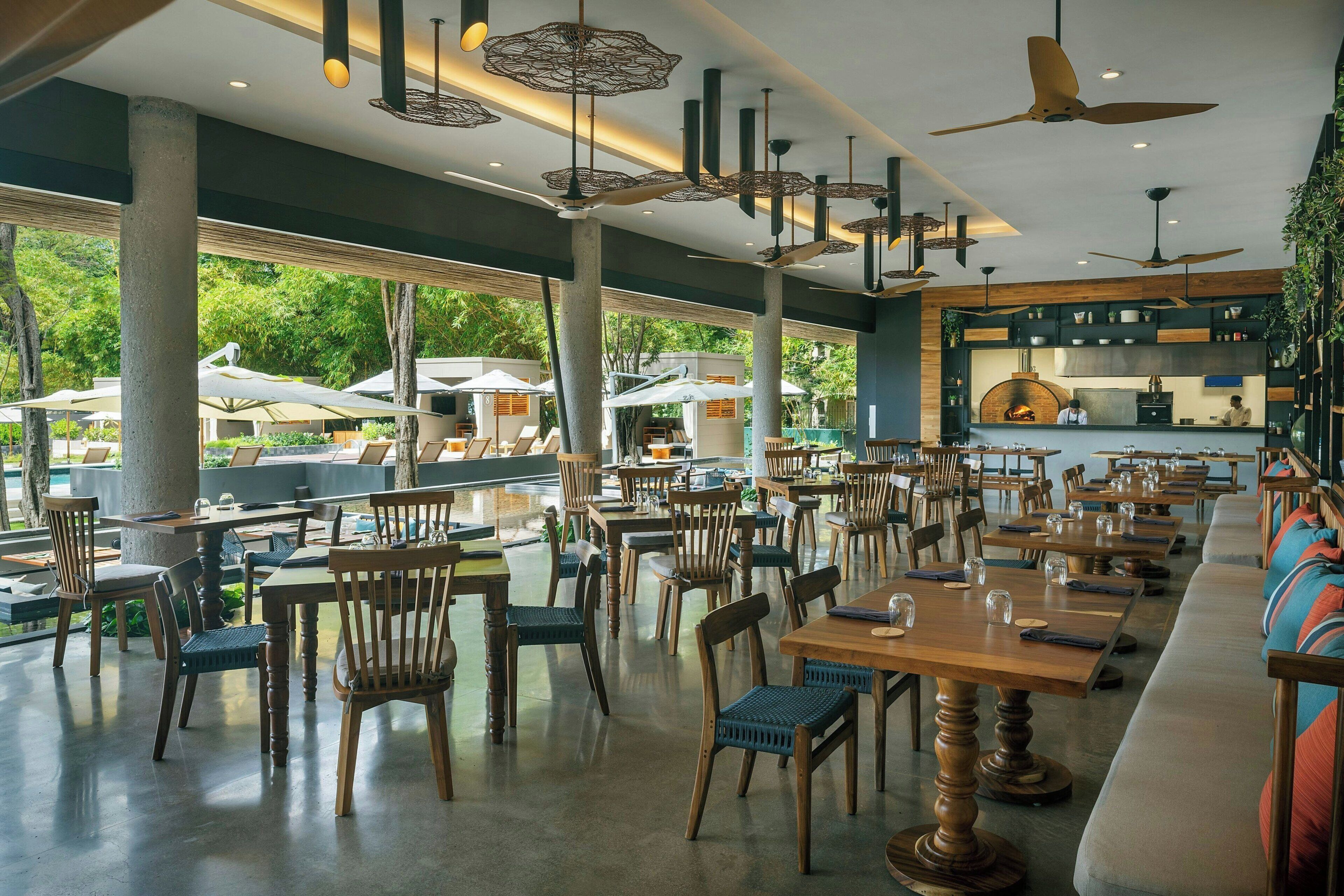 Un restaurant spacieux avec des tables et des chaises en bois, des ventilateurs de plafond et de grandes fenêtres donnant sur un jardin.