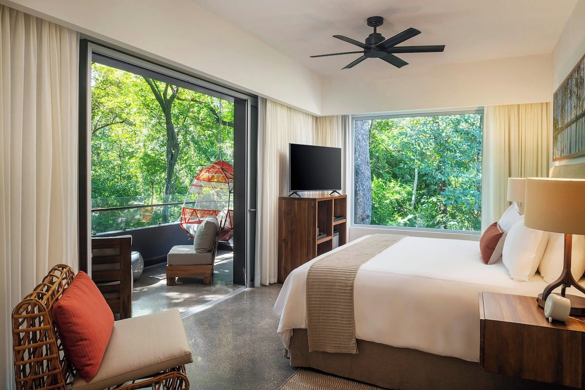 Ein Hotelzimmer mit einem großen Bett, einem Fernseher, einem Balkon mit Stuhl und Tisch sowie einem Deckenventilator.