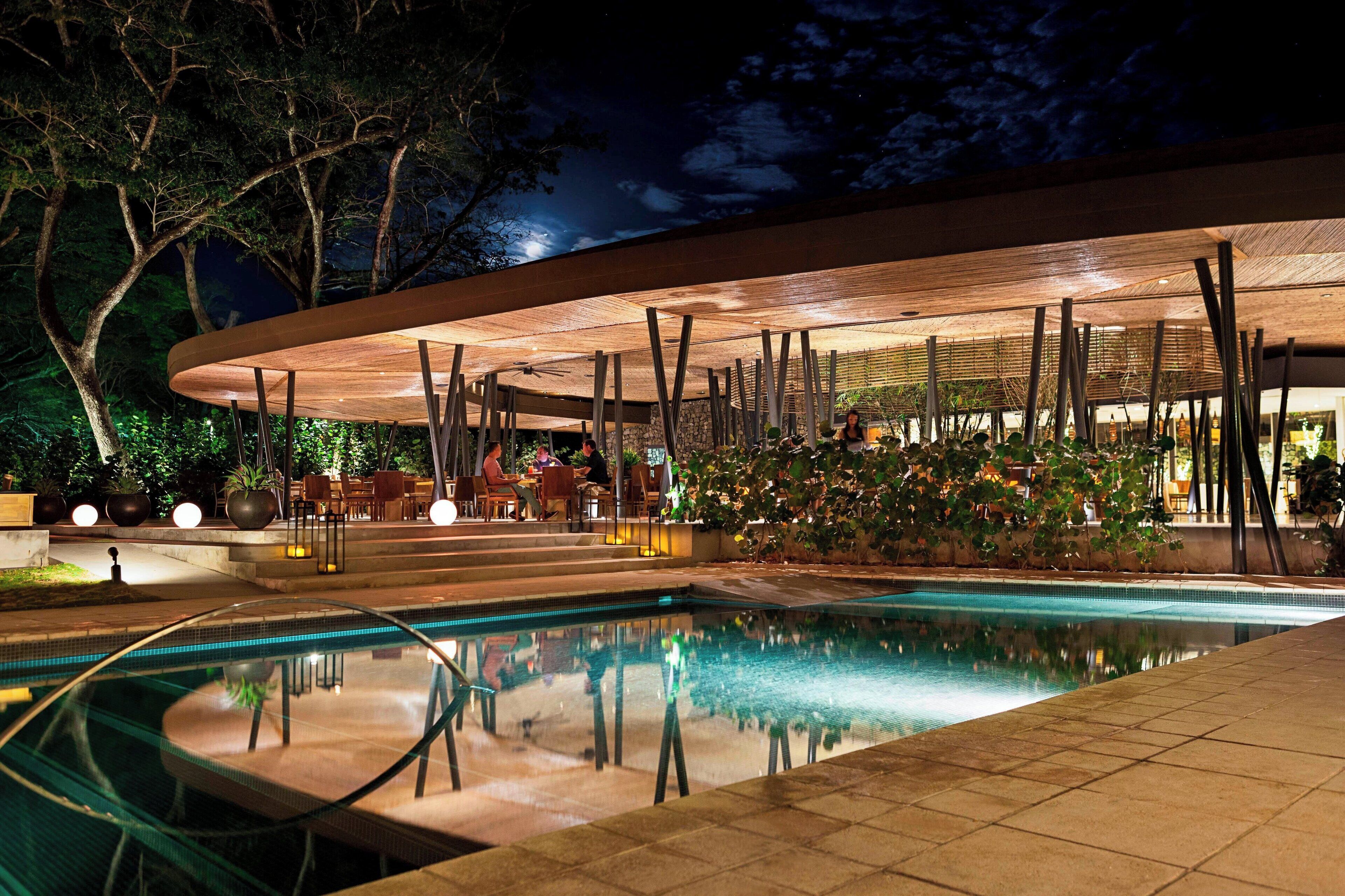 A poolside area with a covered seating area, a pool, and surrounding greenery.