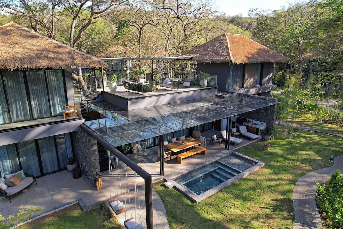 Eine moderne Villa mit einer glasüberdachten Terrasse, Sitzgelegenheiten im Freien, einem Swimmingpool und einem Reetdachgebäude.