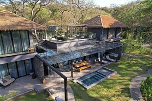 Una villa moderna con un patio de cristal, mobiliario exterior, piscina y una estructura con techo de paja.