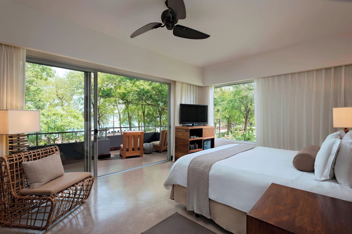 Ein Hotelzimmer mit einem großen Bett, einem Sofa, einem Fernseher und einem Balkon mit Blick auf Bäume.