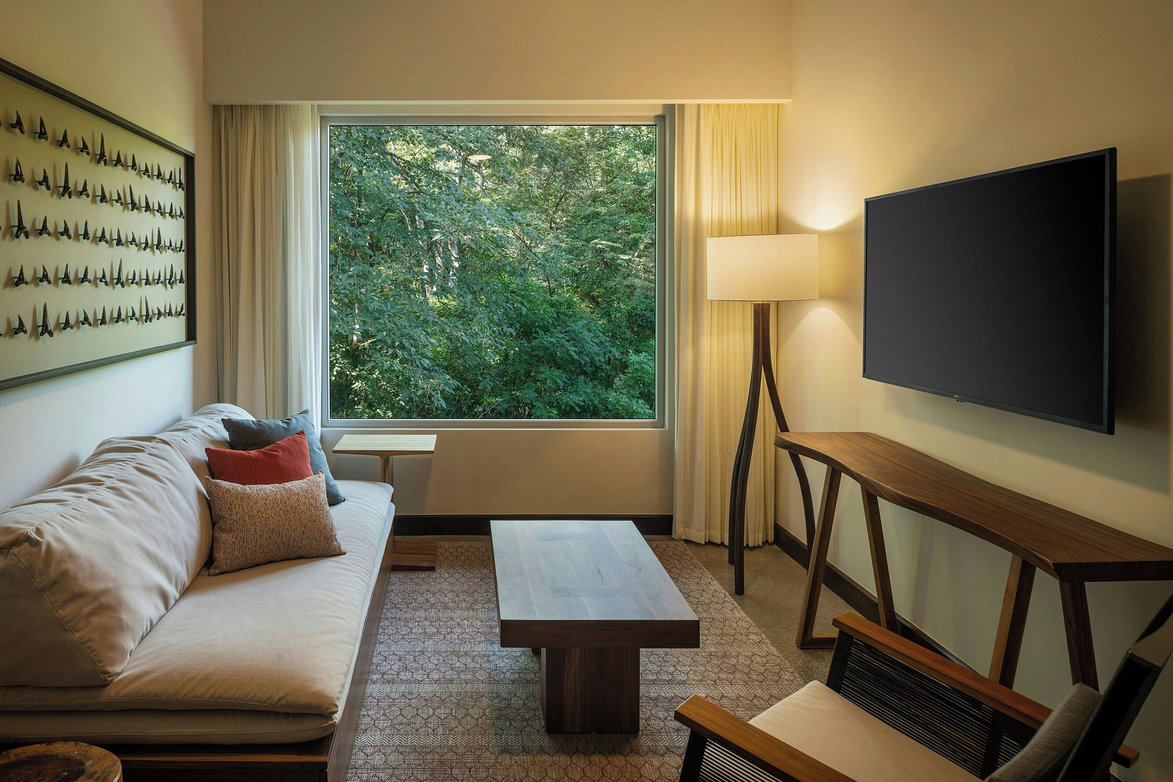 A modern hotel room with a large window, a flat-screen TV, a sofa with cushions, a wooden coffee table, and a lamp.