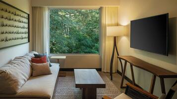 A modern hotel room with a large window, a flat-screen TV, a sofa with cushions, a wooden coffee table, and a lamp.
