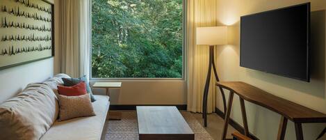 A modern hotel room with a large window, a flat-screen TV, a sofa with cushions, a wooden coffee table, and a lamp.