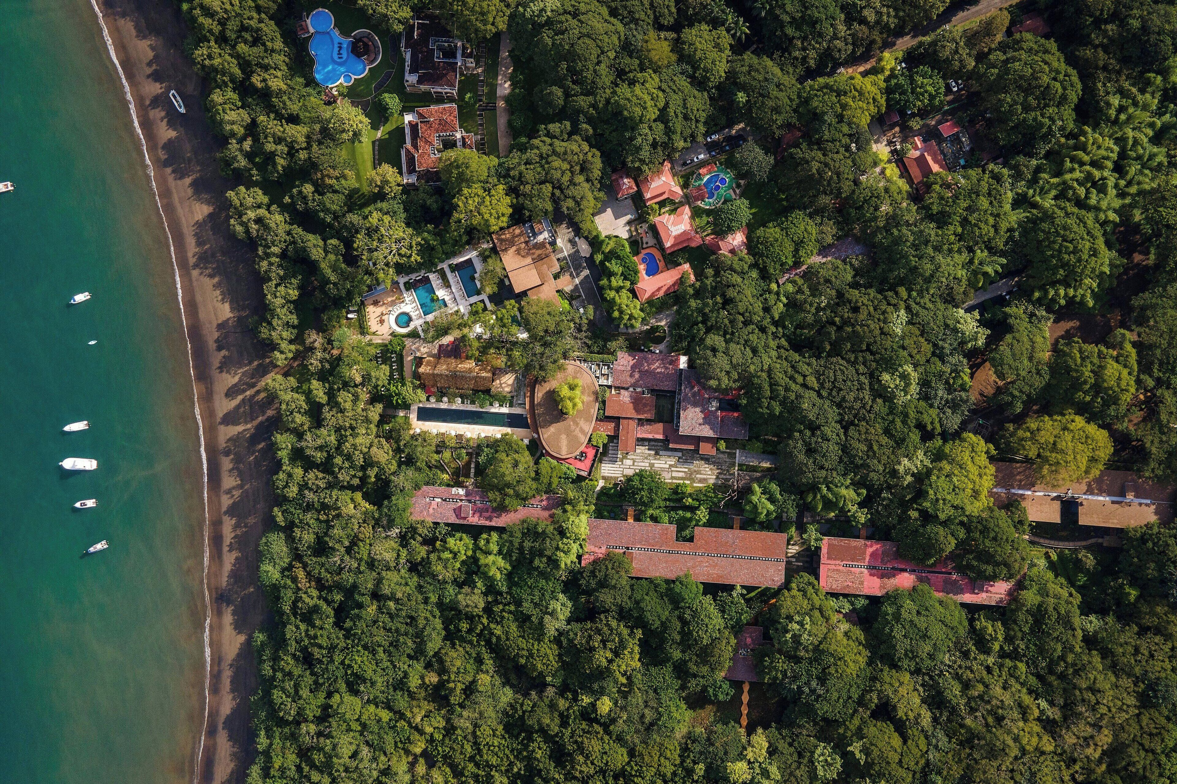 Vista aérea de una propiedad costera con playa, piscina y varios edificios rodeados de vegetación exuberante.