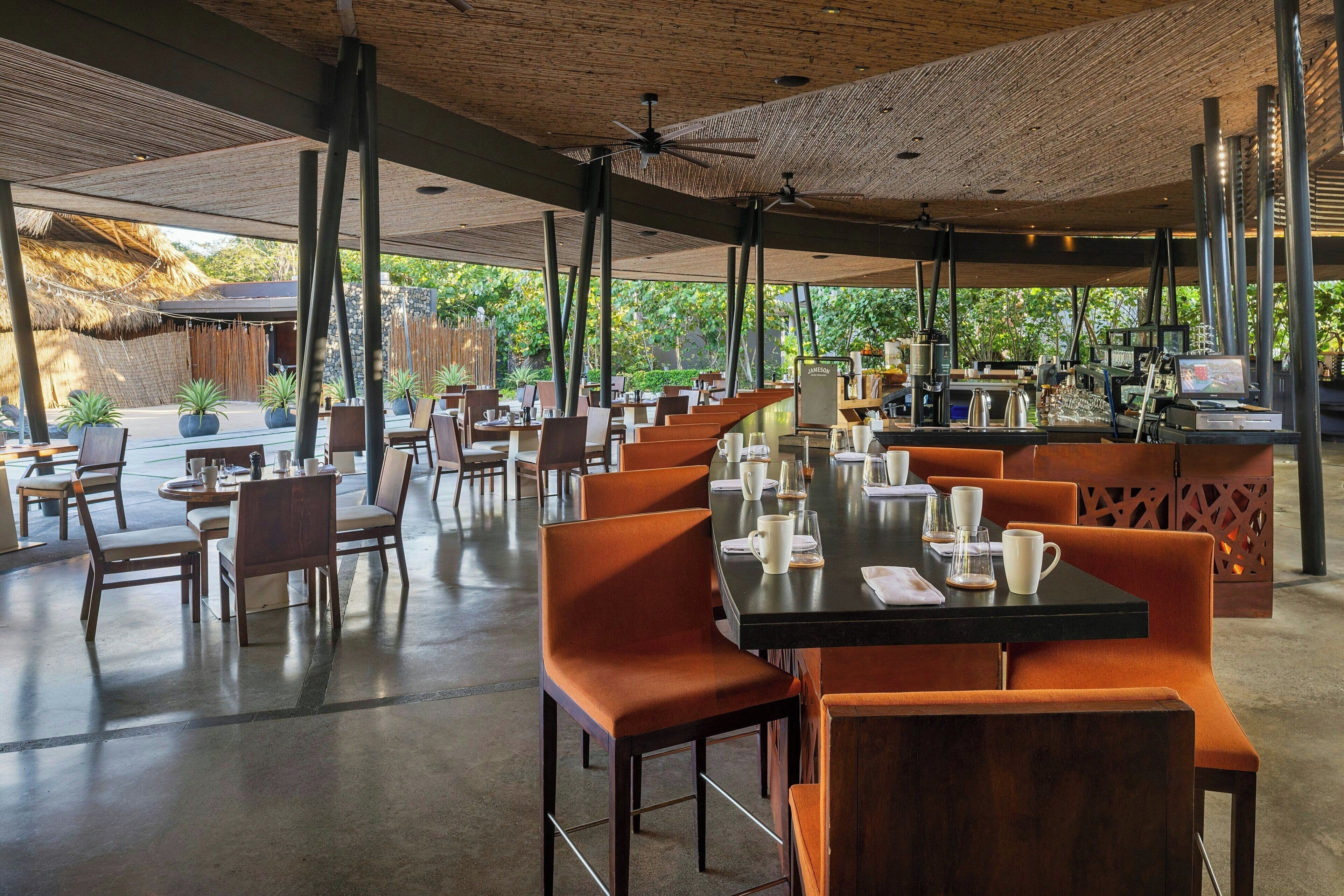 Een ruime eethoek buiten met meerdere tafels en stoelen, een bar en een rieten constructie op de achtergrond.