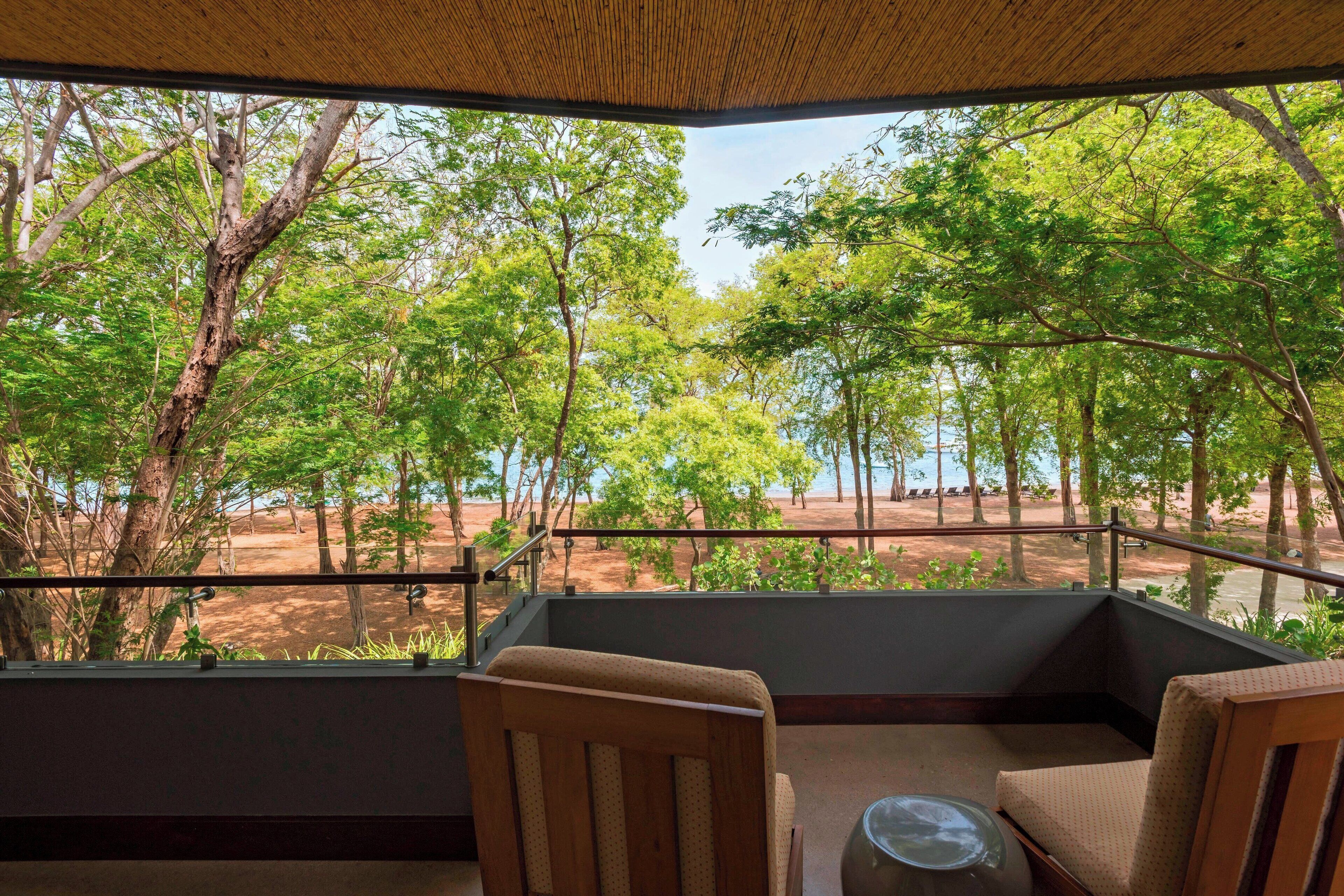 Un balcon avec vue sur une forêt et une plage. On y trouve deux chaises et une petite table.