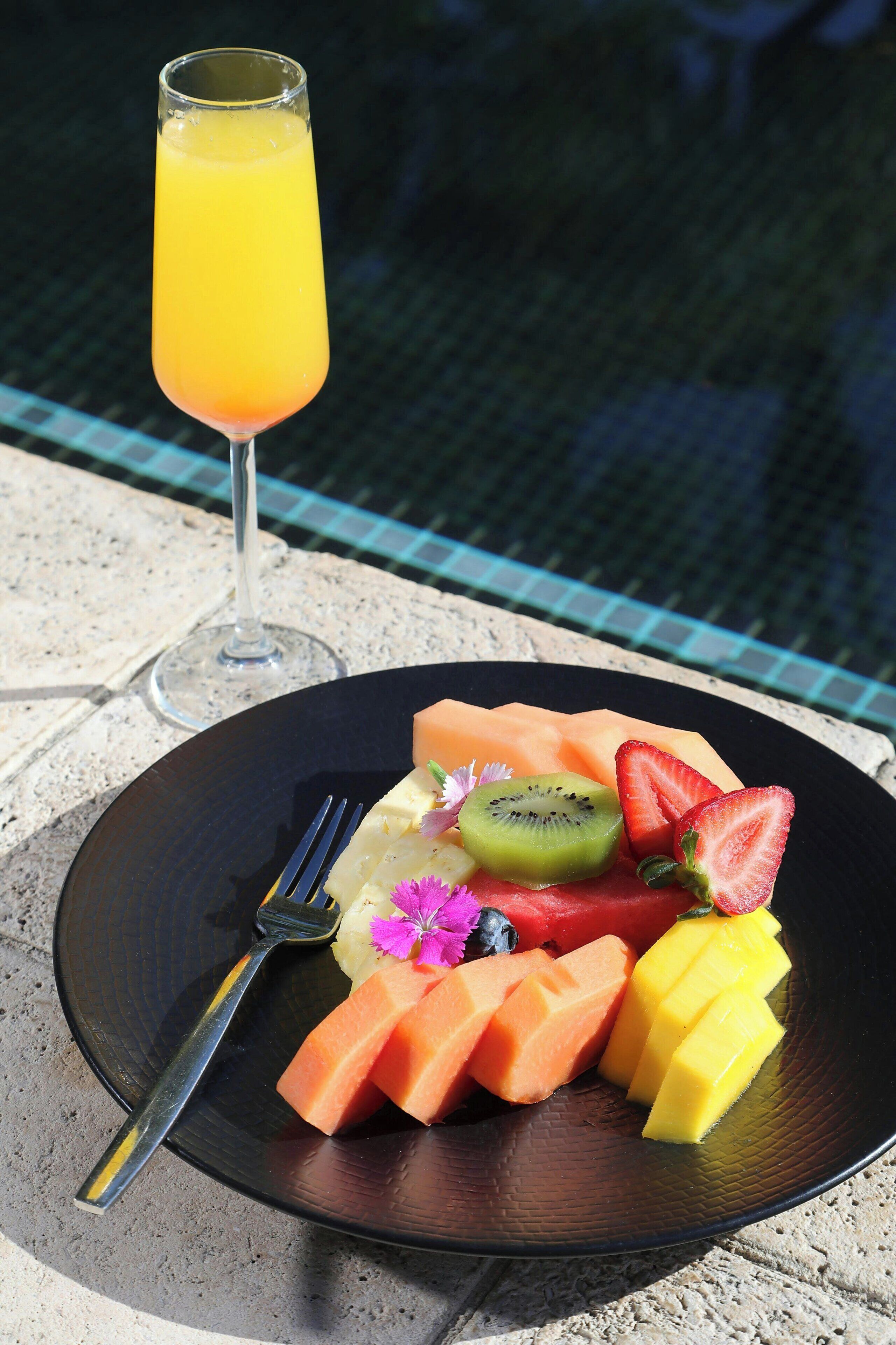 A plate of fruit and a glass of orange juice on a table by a pool.