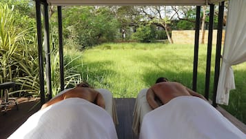 A view from a covered outdoor area with a wooden deck, looking out over a grassy yard with trees and a wooden fence in the background.