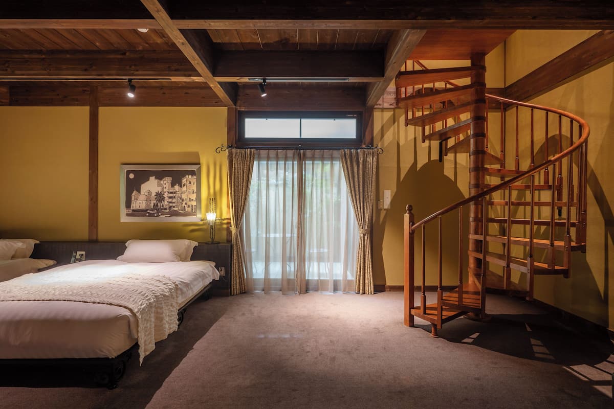 Un dormitorio con una escalera de madera, un ventanal con cortinas translúcidas y una cama con cubrecama blanco.