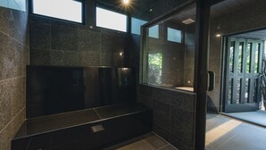 Un baño moderno con baldosas oscuras, un banco y una puerta de vidrio que da a un espacio exterior con vegetación.