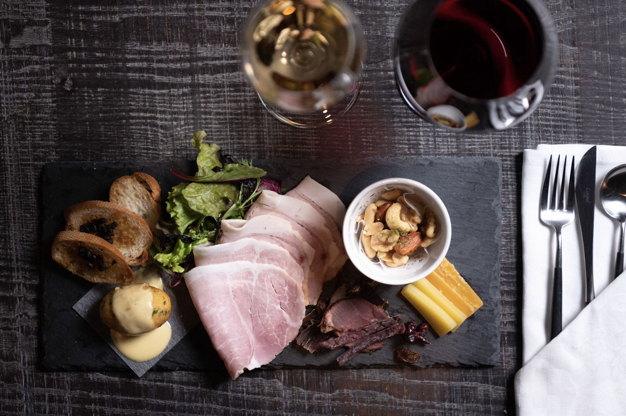 Une planche de charcuterie comprenant différentes sortes de viandes, de fromages et de noix, présentée sur une ardoise, accompagnée d’une bouteille de vin et d’un verre de vin rouge, le tout posé sur une table en bois.
