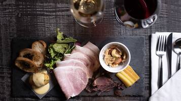 Une planche de charcuterie comprenant différentes sortes de viandes, de fromages et de noix, présentée sur une ardoise, accompagnée d’une bouteille de vin et d’un verre de vin rouge, le tout posé sur une table en bois.