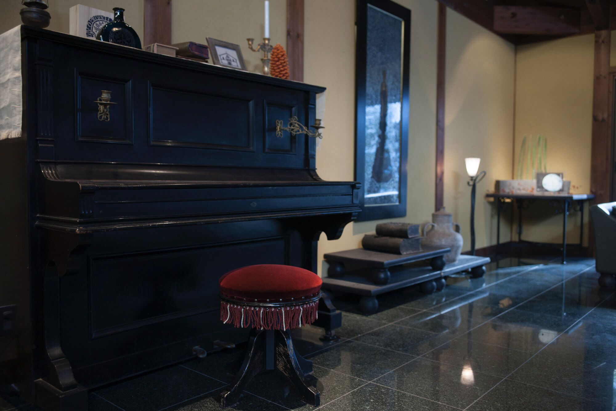 Un piano noir avec un tabouret rouge, dans une pièce dont le sol est recouvert de carreaux, un tableau encadré au mur et une petite table sur laquelle se trouve un vase.