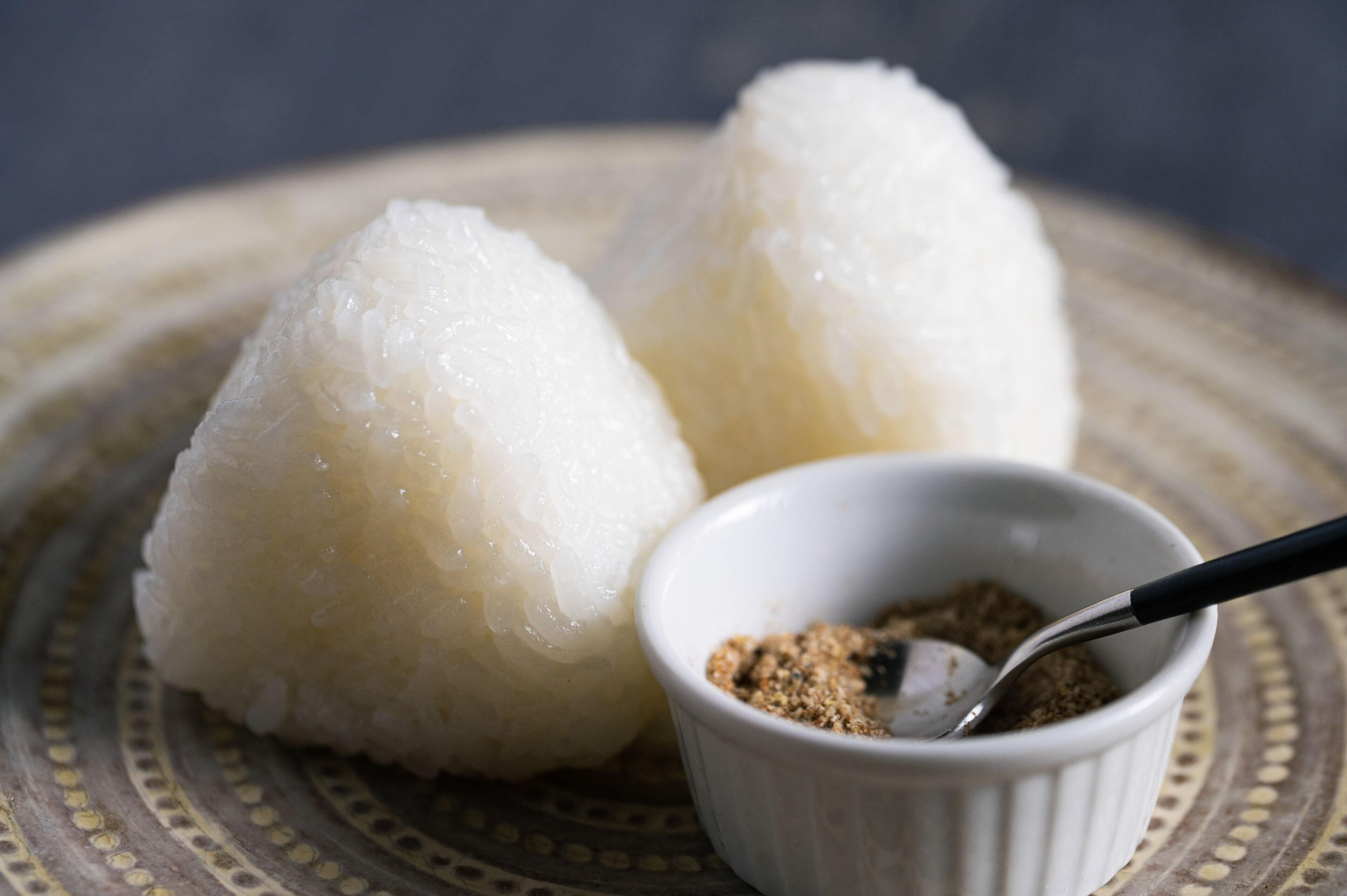 Two white rice balls on a patterned plate with a small bowl of seasoning.