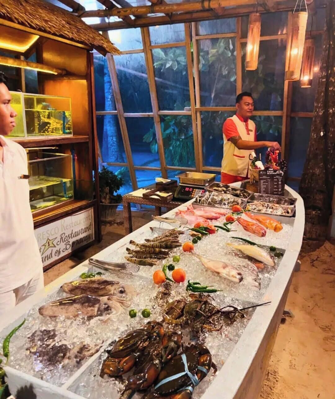 A seafood display with various types of fish and seafood on ice, a counter with a menu, and a rustic interior with a thatched roof.