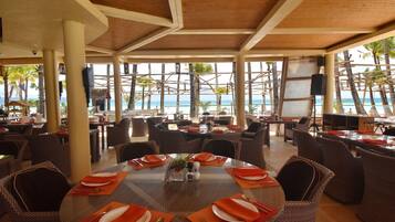 Un restaurante frente a la playa con mesas redondas dispuestas para comer, rodeado de sillas de mimbre y con vista al océano.