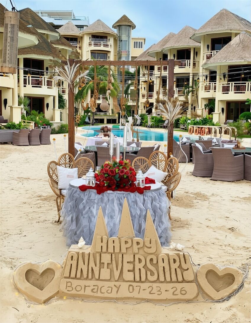 A beachside anniversary setup with a table, chairs, and a sand sculpture.