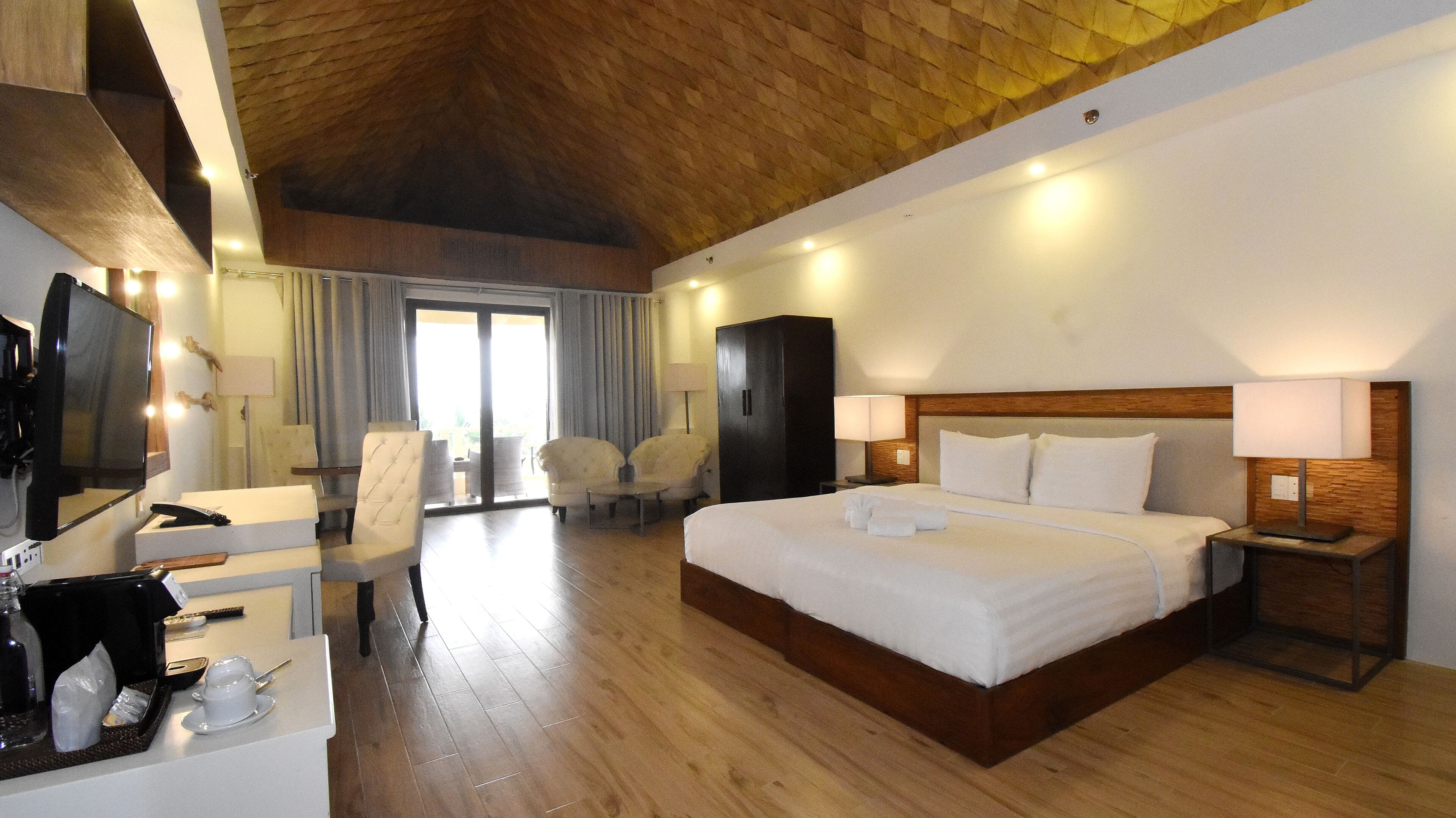 A hotel room with a large bed, a desk with a computer, a TV, and a seating area with chairs.