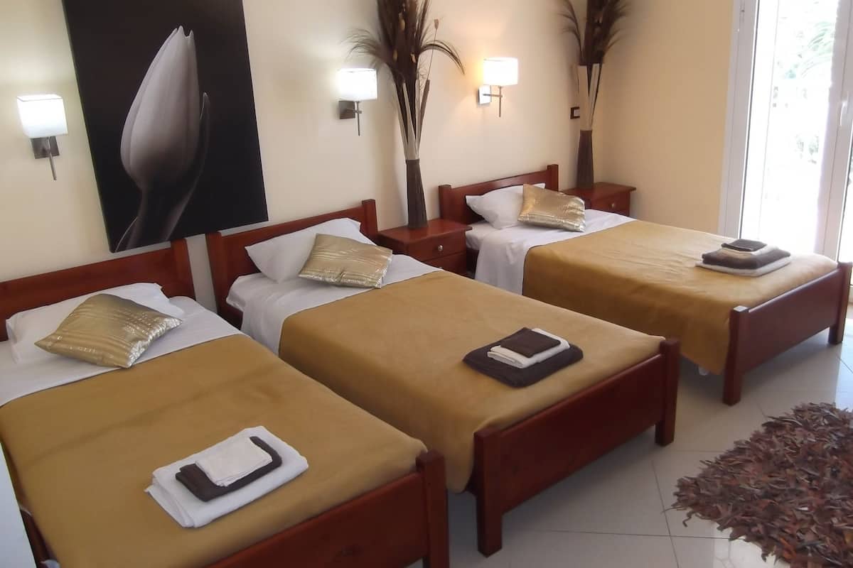 A hotel room with three single beds, each with a brown bedspread and white pillows. There are wall-mounted lamps and a large painting on the wall.