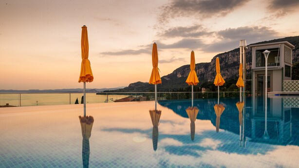 Una piscina infinita con agua cristalina que refleja el cielo y las montañas, rodeada de sombrillas amarillas y un edificio junto a la piscina.