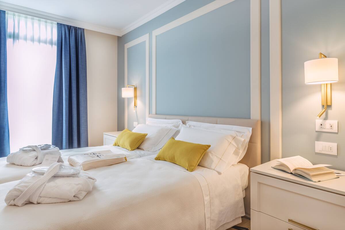 A hotel room with two beds, a bedside table with a lamp, a bedside table with a book, and a window with curtains.