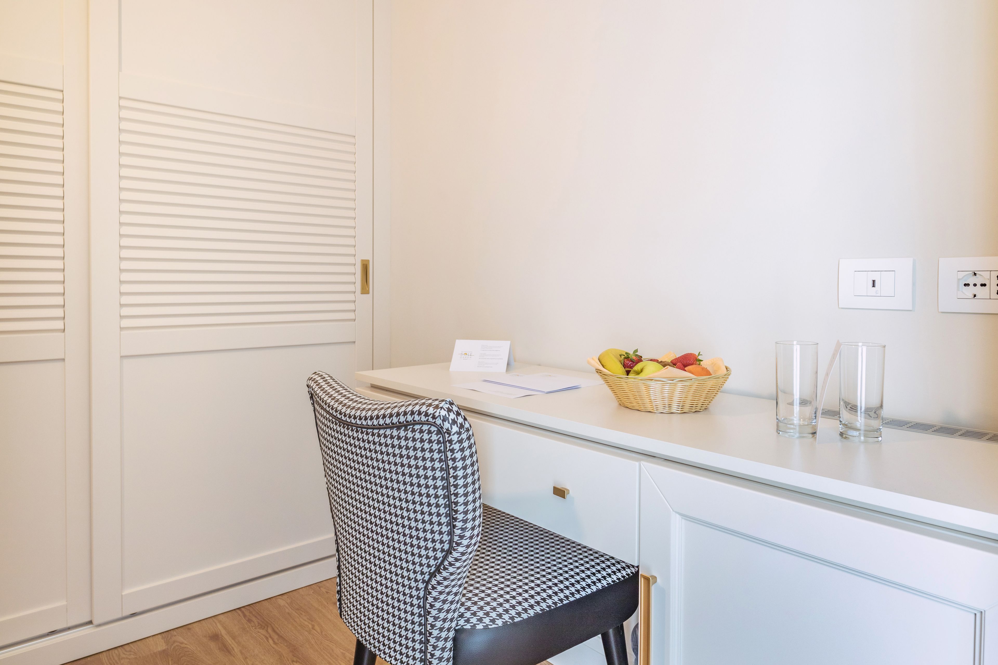 A room with a white desk, a houndstooth chair, a basket of fruit, and two glasses.