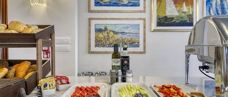 Una mesa de buffet con diversos alimentos y salsas, incluyendo tomates en rodajas, pepinos y pimientos, además de pan y pasteles.