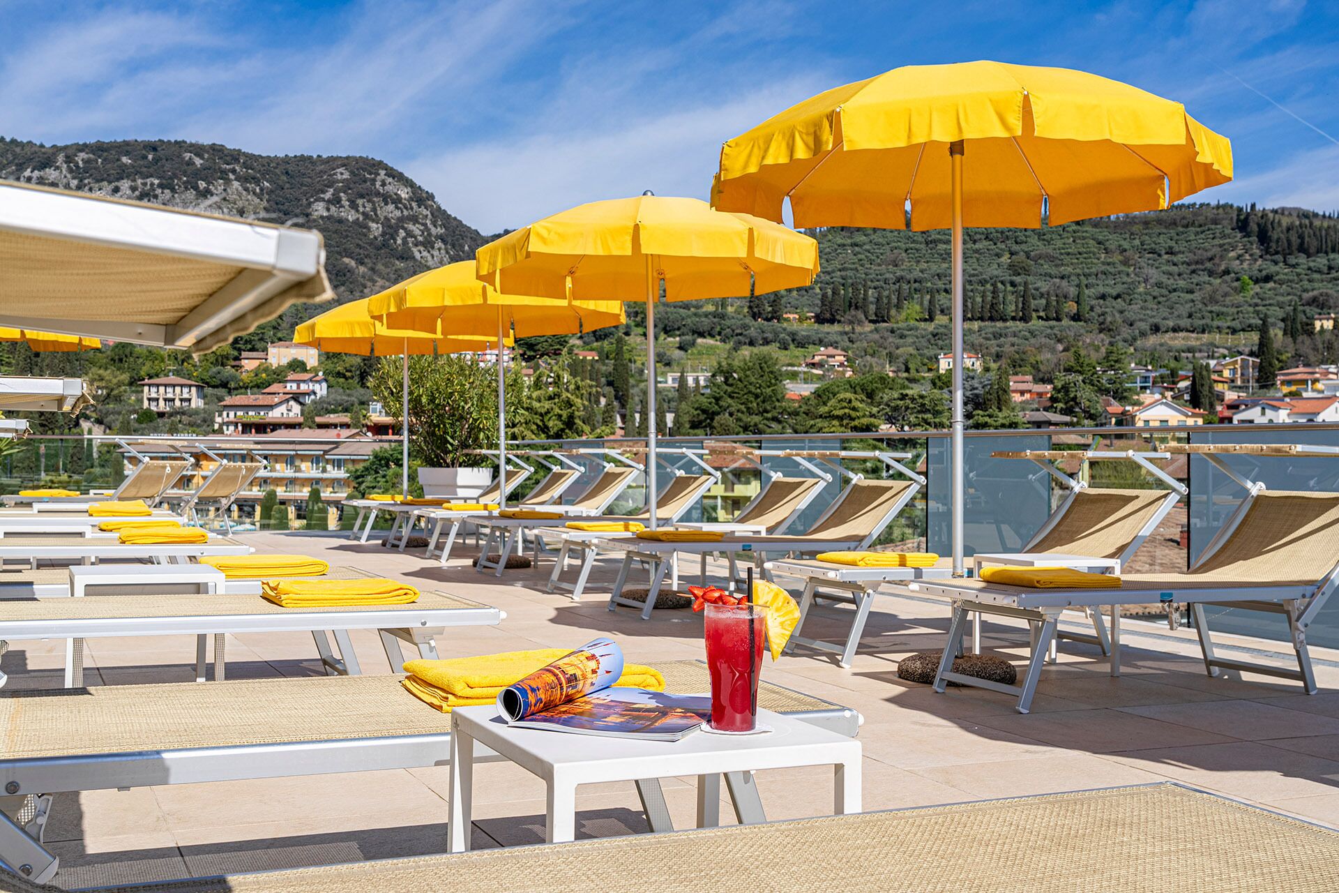 A rooftop pool area with sun loungers, yellow umbrellas, and a table with a drink and a magazine.